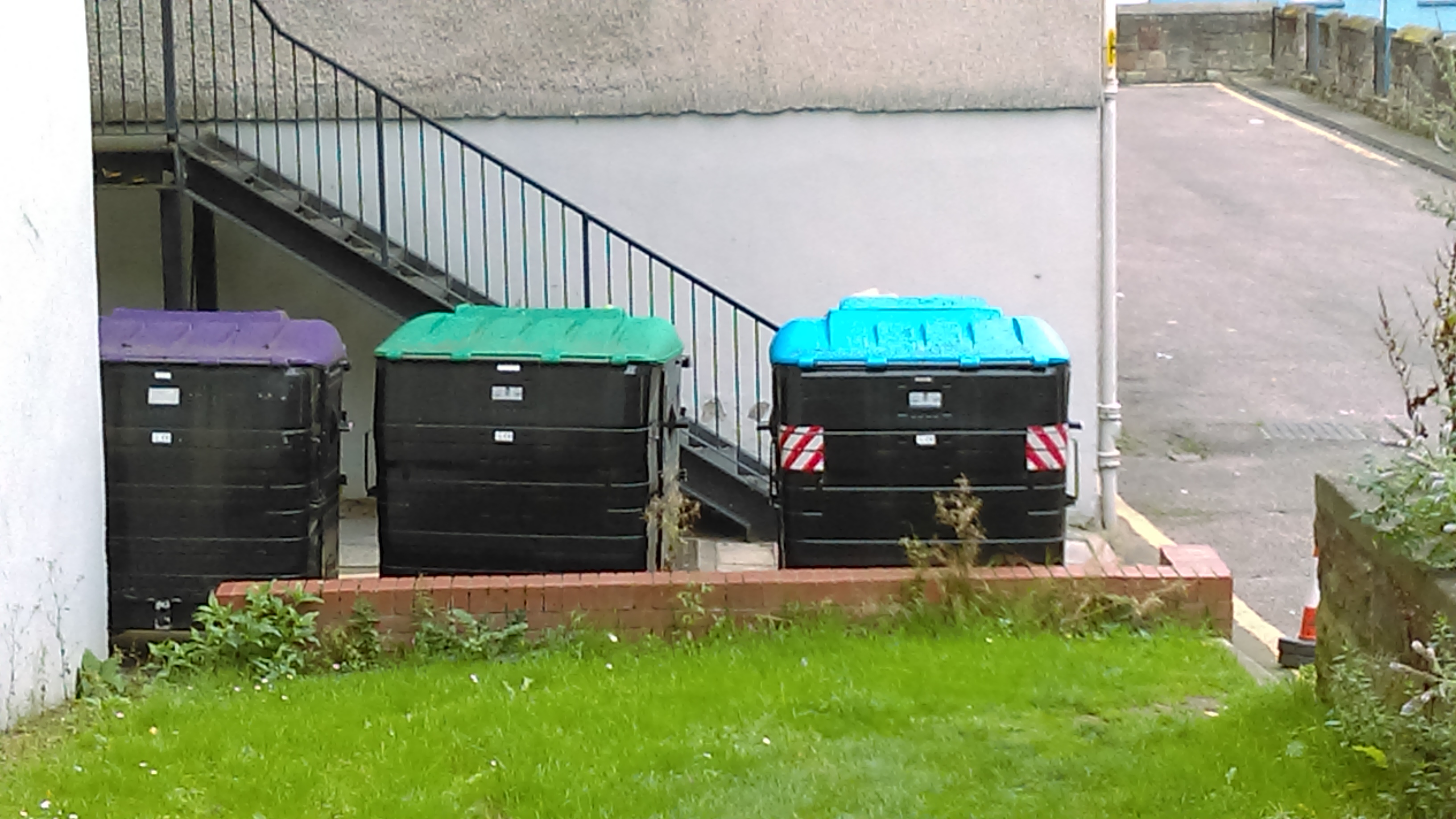Tres contenidors de reciclatge, un lila per al vidre, un verd per reciclatge mixt i un de blau per al paper i cartró